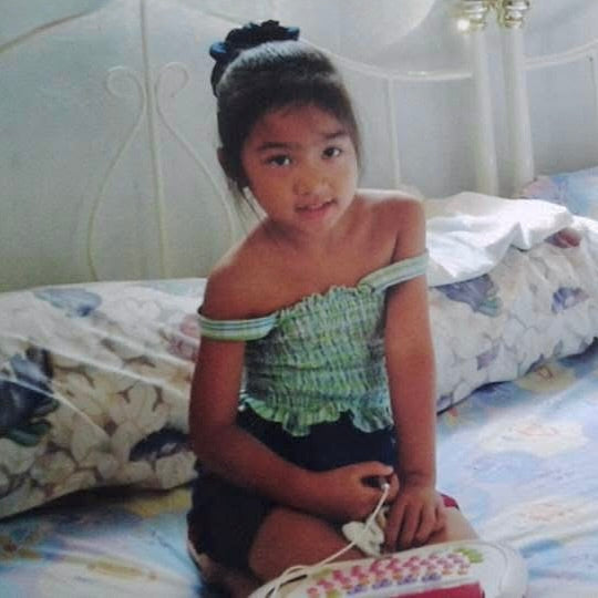 Young Jessa sitting on a bed wearing a green off-shoulder top in a bedroom setting.